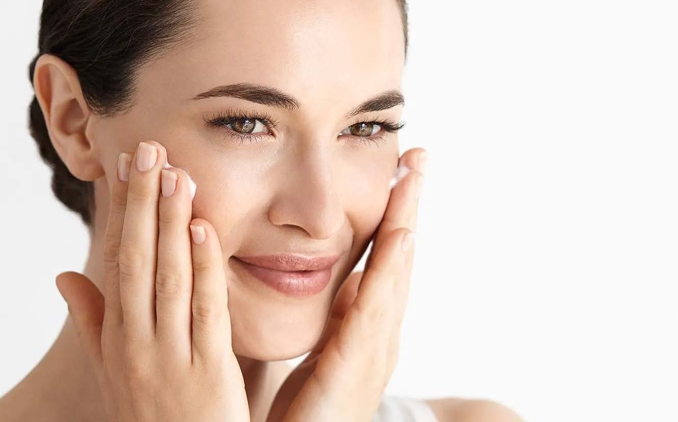 beautiful woman skin complexation hands on face rubbing cream into cheeks close-up subtle smile brown eyes dark hair